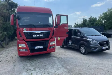 Czerwona ciężarówka MAN z otwartymi drzwiami obok ciemnego vana Ford.
